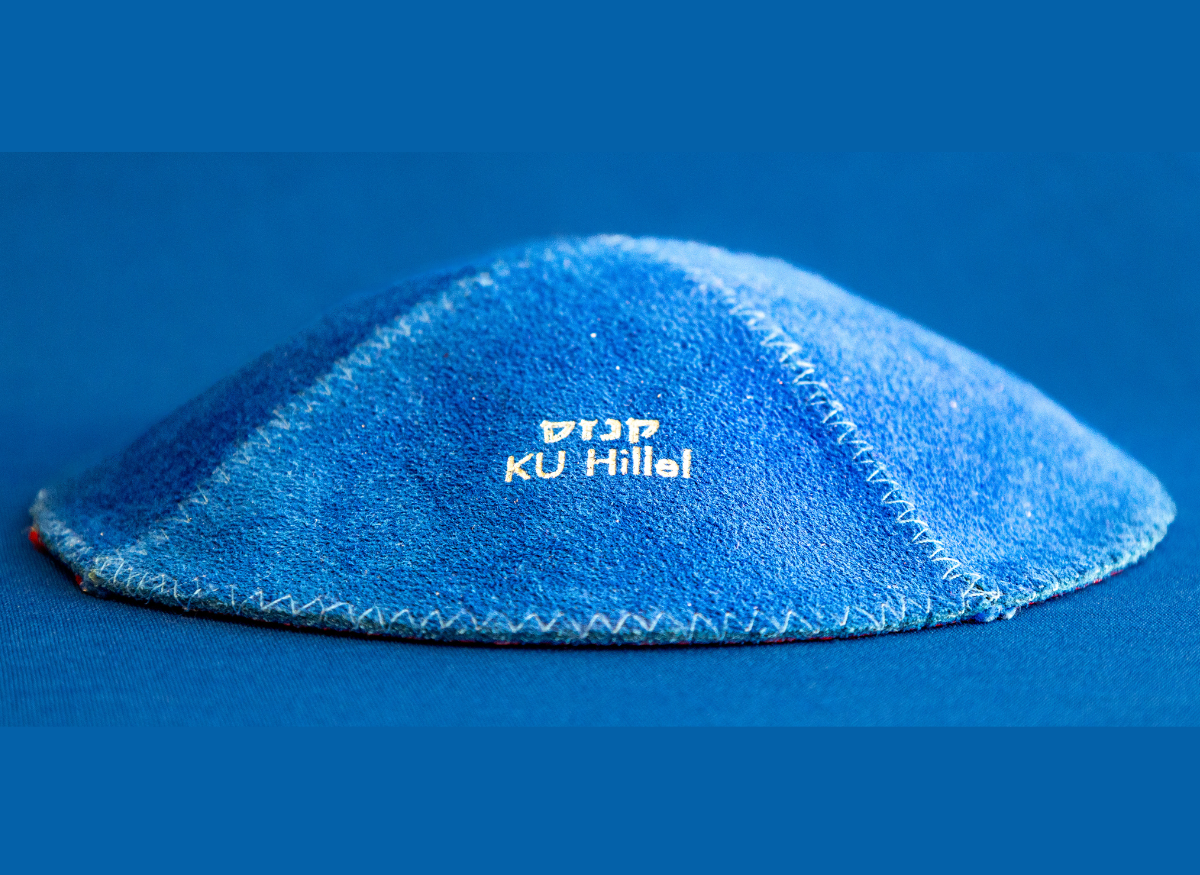 A blue kippah with white embroidered text reading KU Hillel 